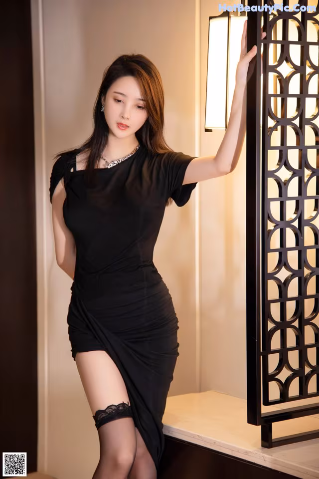 A woman in a black dress leaning against a wall.