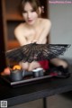 A naked woman sitting at a table holding a black fan.